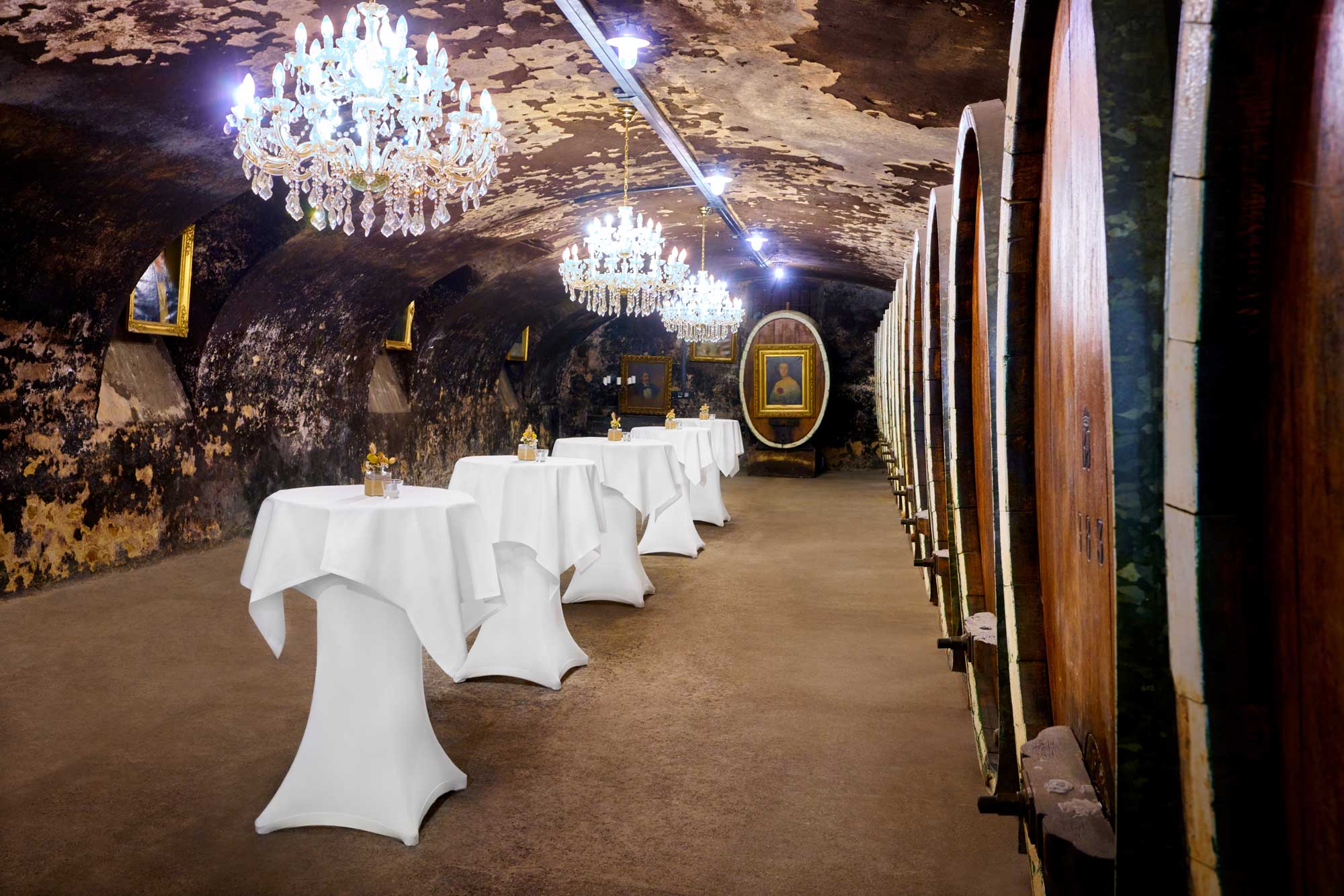 Historischer Fasskeller "Cave 1822". Das geschichtsträchtige Kellergewölbe im Anwesen Ketschauer Hof wird von prunkvollen Kristallkronleuchtern in Szene gesetzt. Auf der einen Raumseite sind 10 historische Weinfässer aneinander aufgereiht. Linksseitig schmücken Gemälde mit goldverschnörkelten Rahmungen die Nischen aus. Mittig laden 5 Stehtische zum Sektempfang einer Feierlichkeit ein.