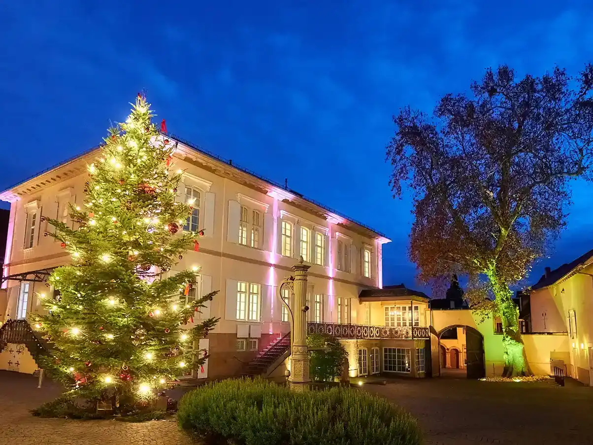 Weihnachtstimmung im Genussresort Ketschauer Hof in Deidesheim, Pfalz. Der Betrachter steht im Innenhof des Anwesens, und blickt auf die angeleuchtete Fassade des Hotelgebäudes sowie auf den 10 Meter hohen, prunkvoll geschmückten Tannenbaum.