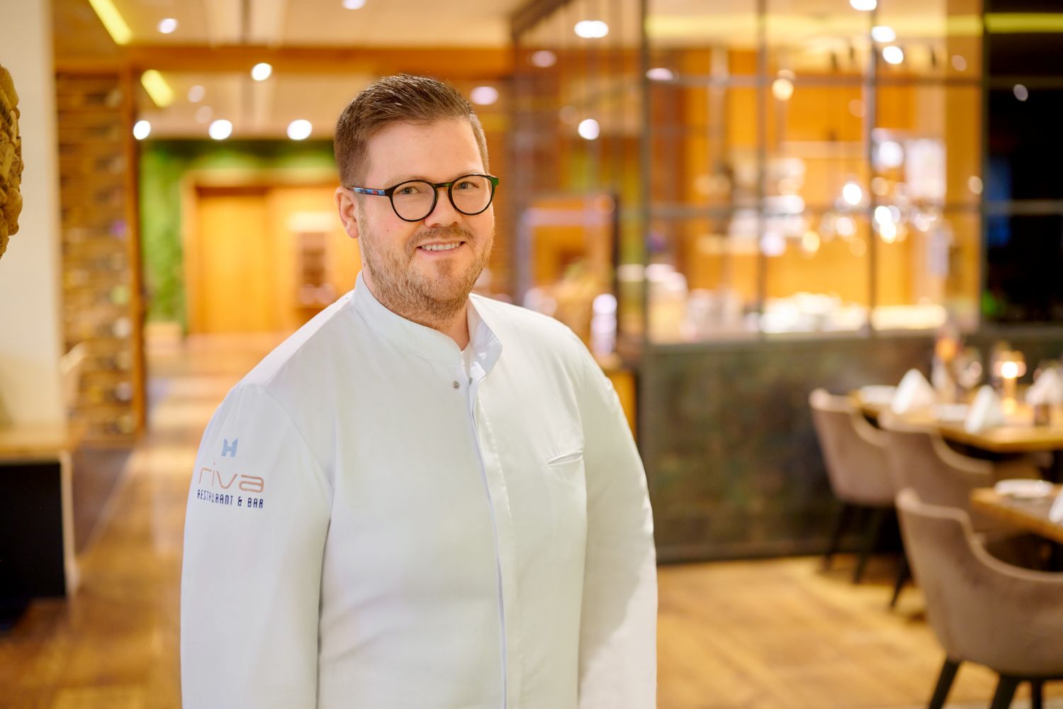 Mike Deuschel, Küchenchef im Restaurant RIVA des Wein- und Seminarhotels Kaisergarten Deidesheim/ Pfalz. Mike Deuschel verantwortet zudem das Restaurant 1718, im Genussresort Ketschauer Hof. Hier, mit Blick auf die offene Showküche im Hintergrund.